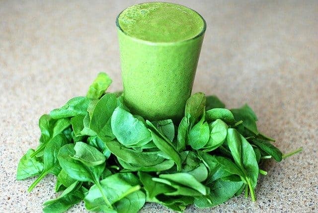 Un bicchiere con un frullato verde, foglie di spinaci tutto intorno